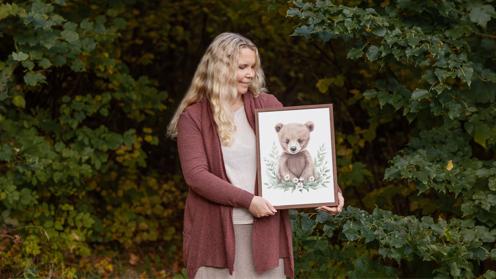Konstnären bakom Woodland Wonders håller en handmålad barntavla i akvarell på björnen Benji, framför ett skogsparti
