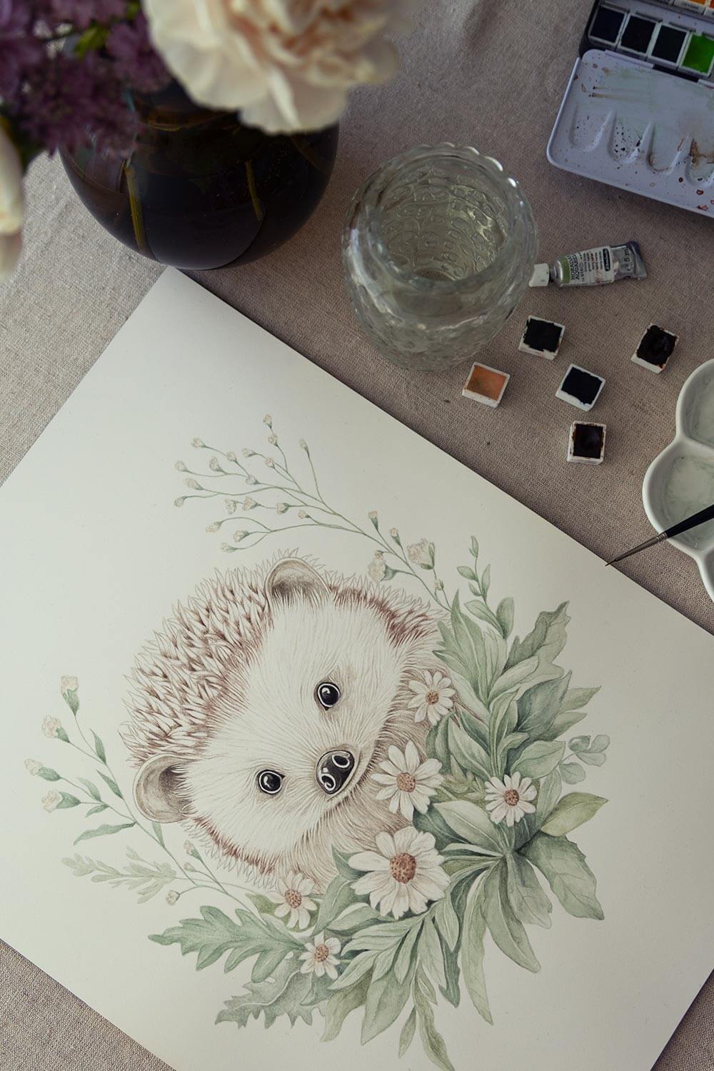 Close-up of a hand-painted watercolor print featuring Nilo the hedgehog and paintbrushes